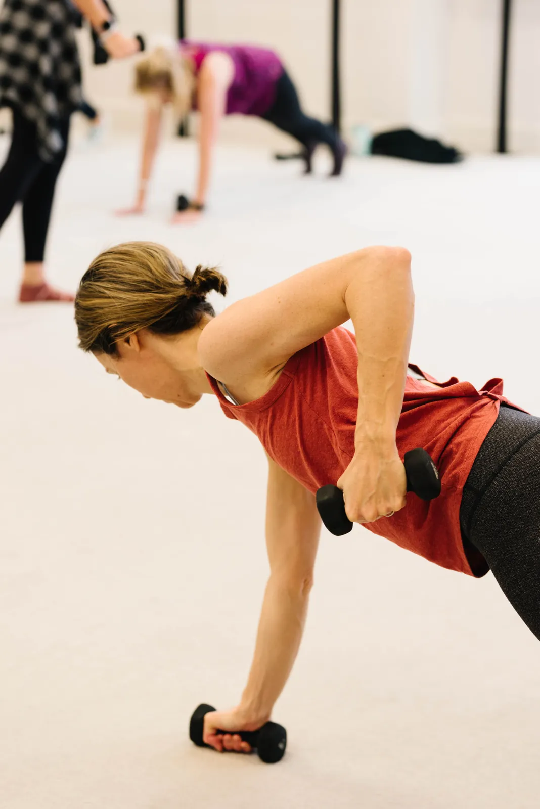 A client rowing a hand weight from a plank position on the studio carpet